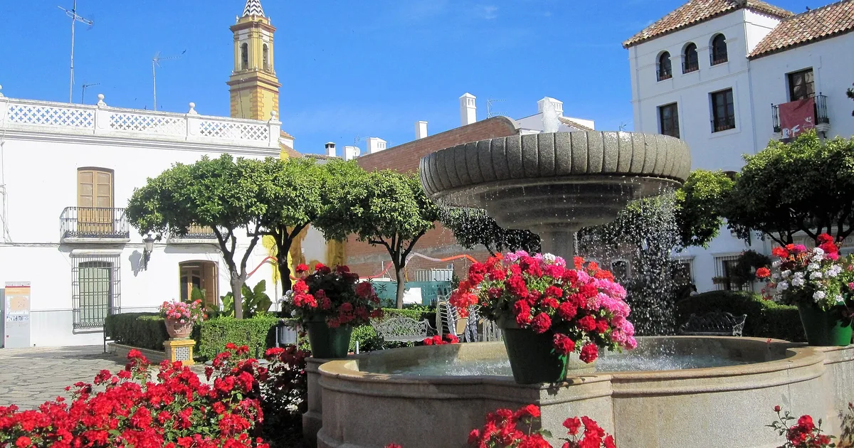 Fountain and red flower beds on the Plaza de las Flores in Estepona old town, with whitewashed houses and a church tower