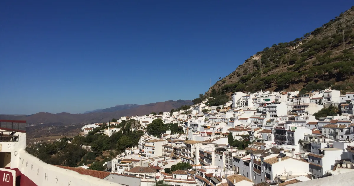 Whitewashed houses of Mijas Pueblo climbing the green hillside of the Sierra de Mijas above the Costa del Sol, with the village bullring wall in the foreground — Mijas Pueblo escorts guide 2026